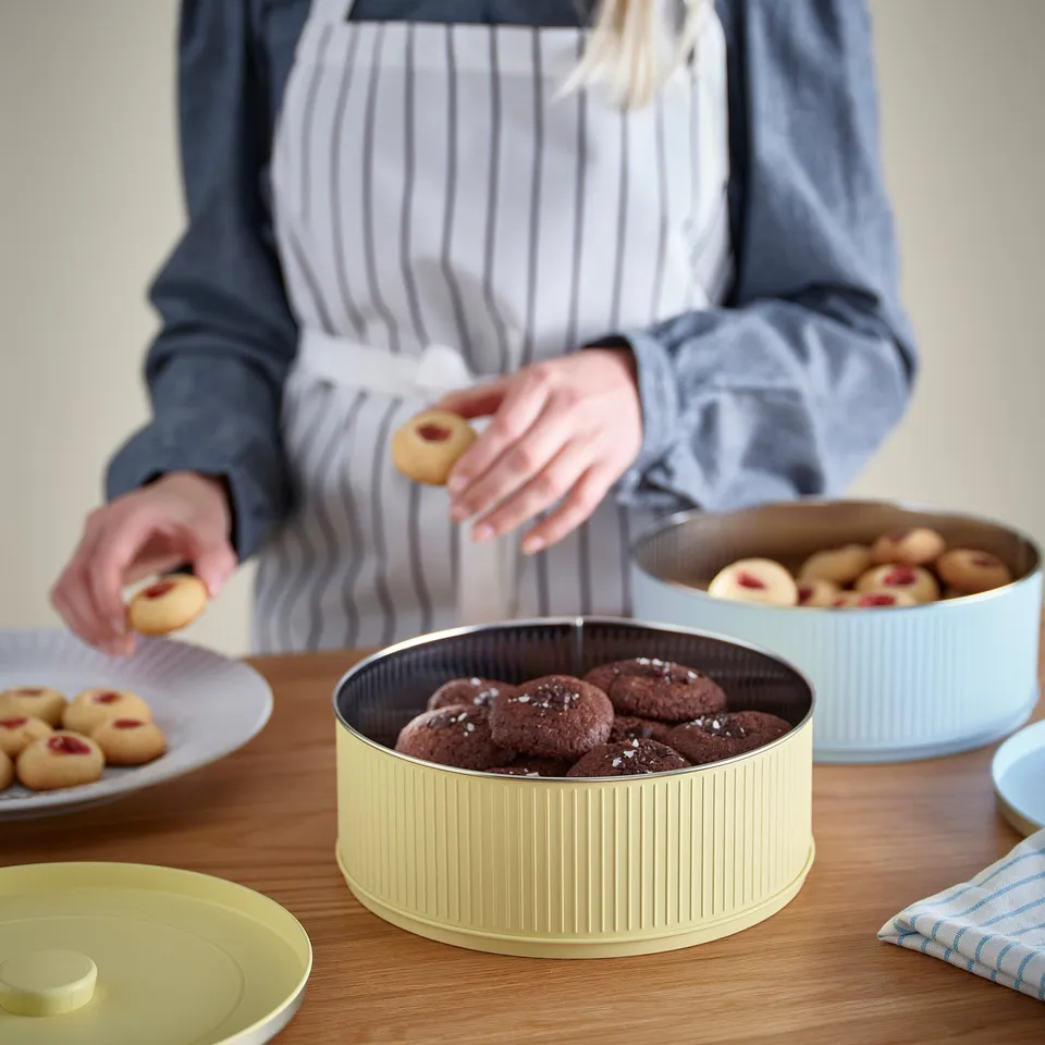Person in apron organising cookies in stylish, pastel-colored BETTHAJ tins on a wooden table.