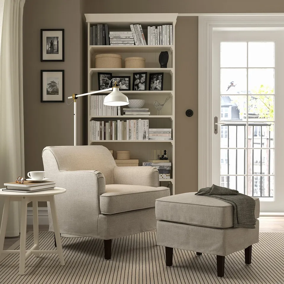Cosy reading nook with beige ROCKSJÖN armchair, matching ottoman, bookshelf & windows.