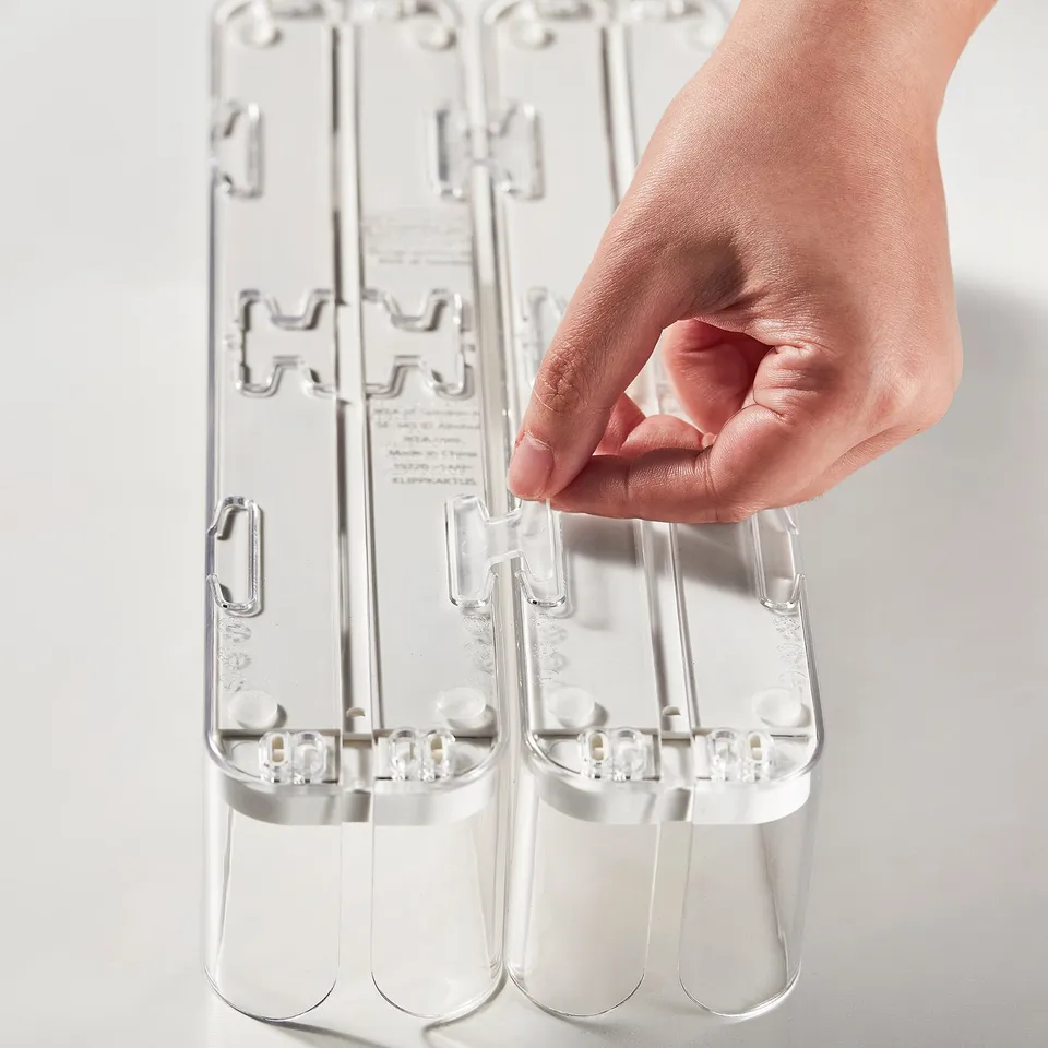Hand holding a transparent, stackable fridge organiser with adjustable sides for cans/bottles.