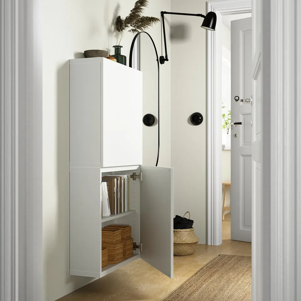White wall-mounted cabinet, open showing shelves with items. Black handles, adjustable shelf, and wall lamp.