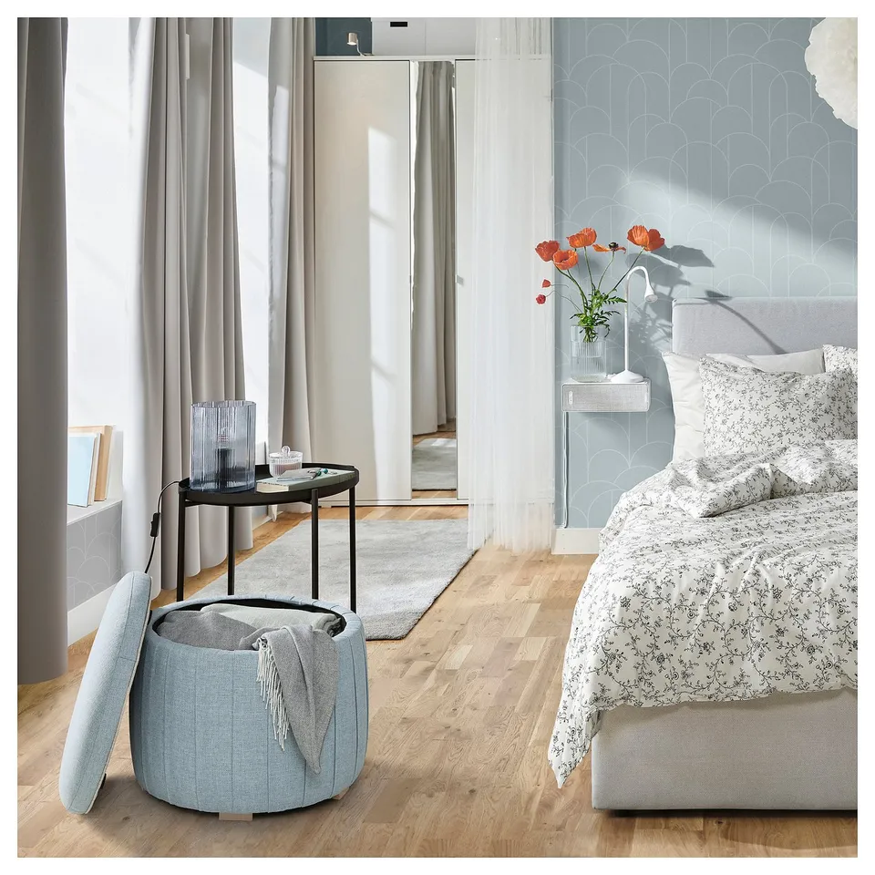 Bright bedroom with wooden floor; white bed, floral duvet; round grey ottoman with lid open revealing storage; small black side table with decoration.