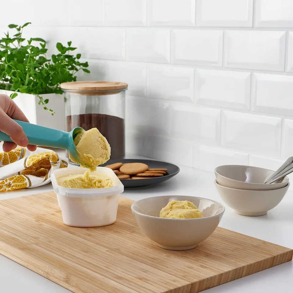 Scooping yellow ice cream into a bowl with a blue scoop; bowls, cookies, jar in background.