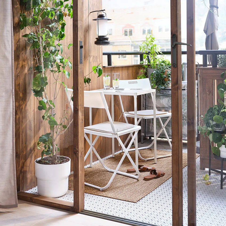 Bright balcony with white FEJAN folding chair, glass table, and lush plants. Wooden floor, natural light, and city view.