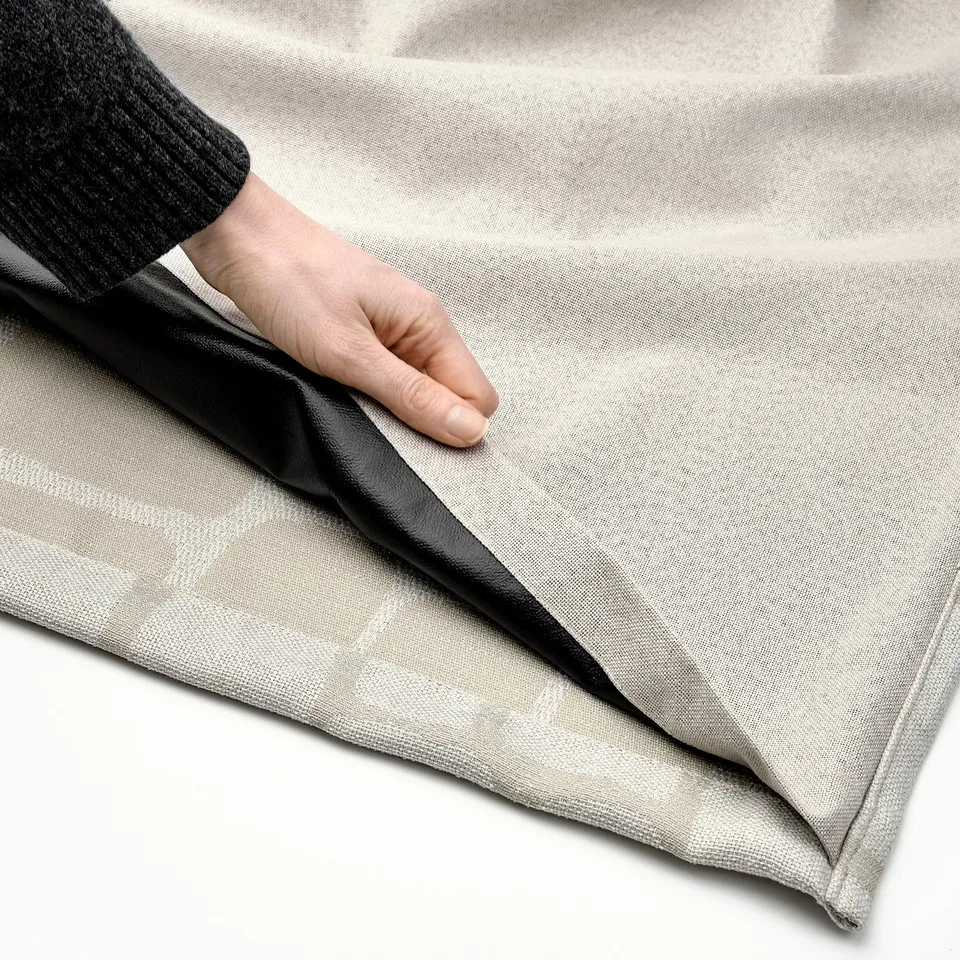 A hand adjusts a beige jacquard LÖNNSTÄVMAL block-out curtain, showcasing its elegant texture and light-blocking functionality.
