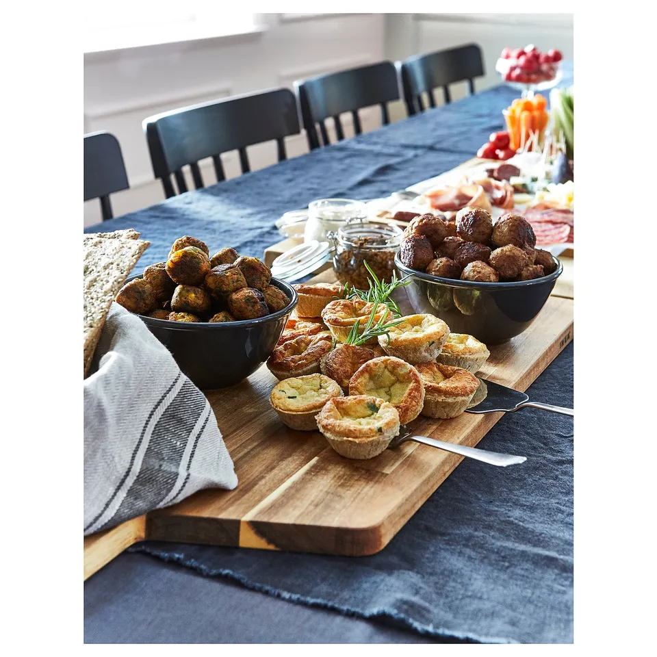 Dark wooden acacia cutting board with varied finger foods and black bowls on blue tablecloth.