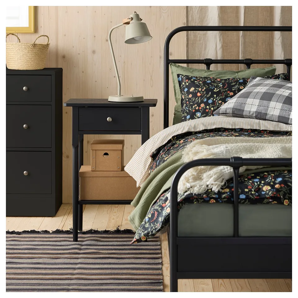 Cosy bed with black frame, floral/checked bedding, green blanket, bedside table, basket, striped rug on wood floor.