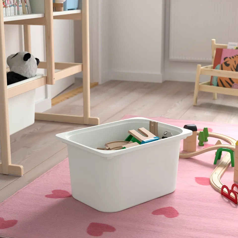 Playroom with light pink rug, white storage bin holding wooden toys. Wooden bookshelf, toy chair, and track nearby.