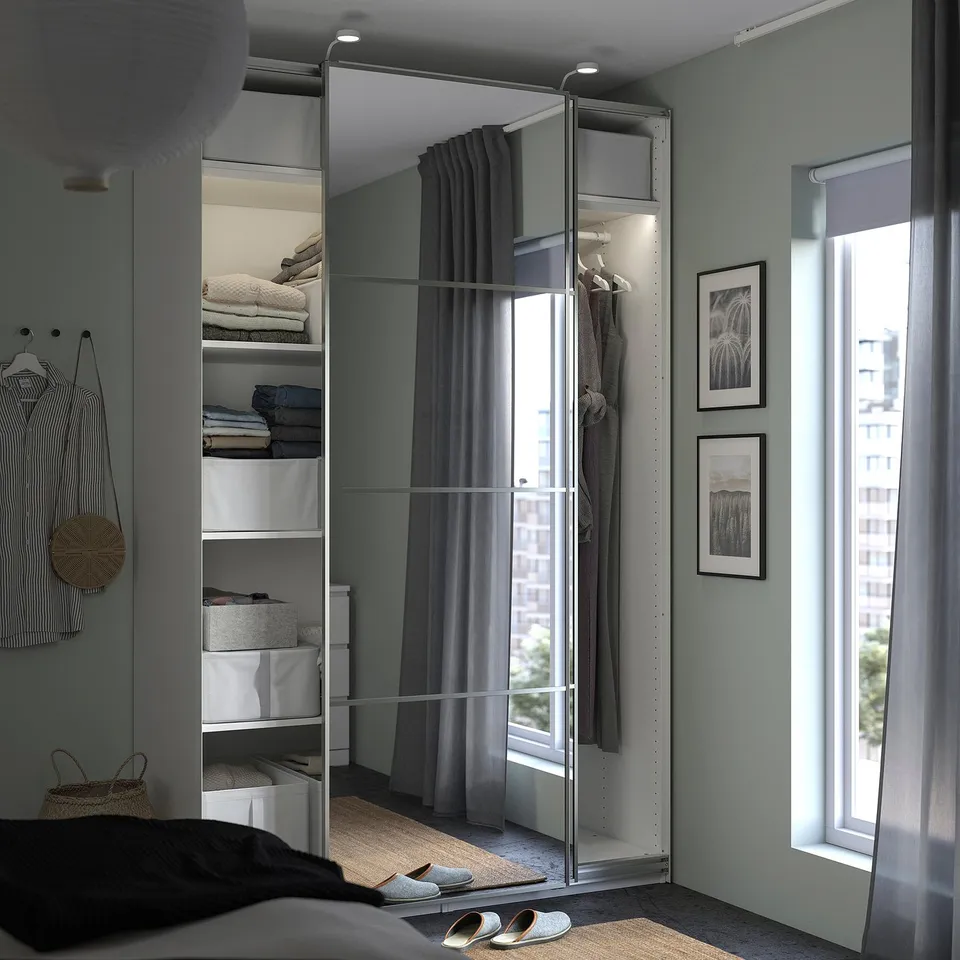 Bedroom with PAX wardrobe in white, mirrored doors, neatly organised clothes, and a window with light curtains.