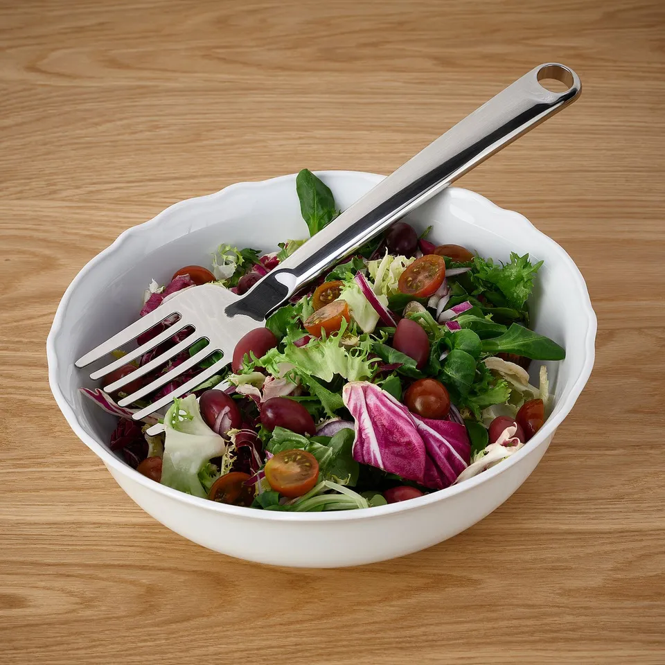 Salad bowl with stainless steel FINMAT fork on wooden table. Contains greens, tomatoes, cabbage.