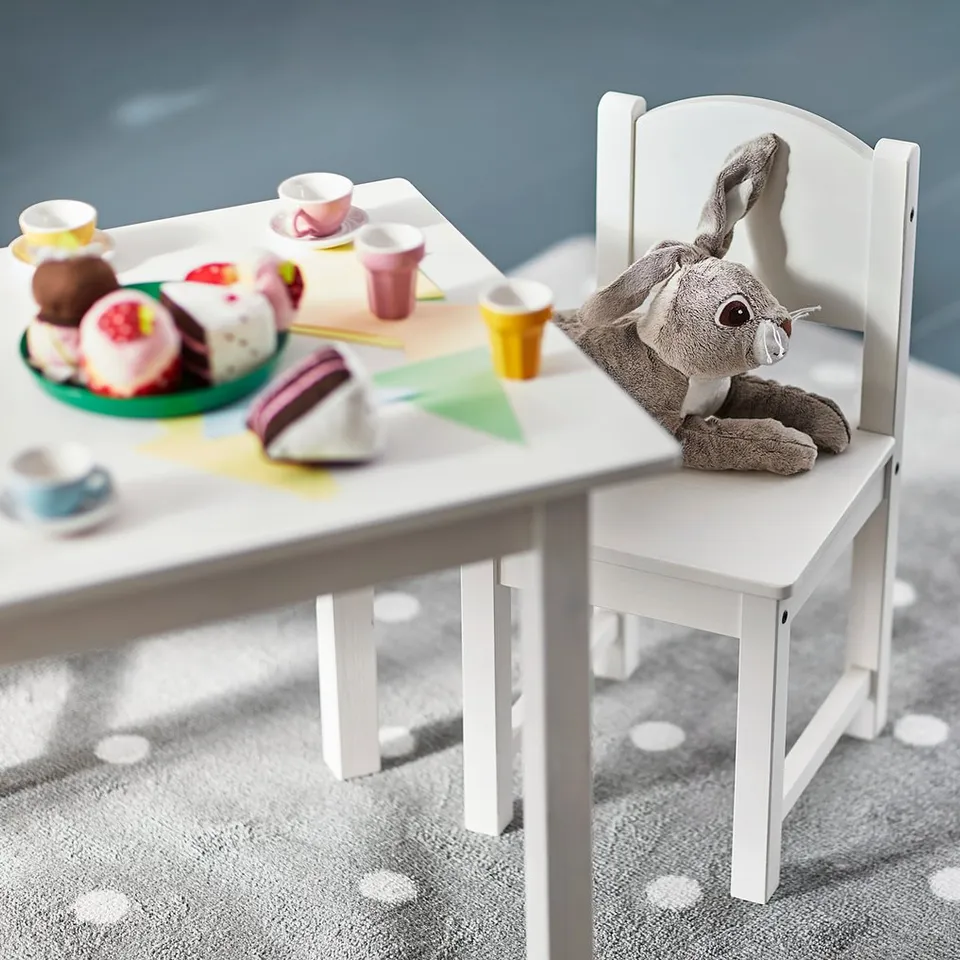 Small white toy table and chair set, perfect for kids play. Stuffed bear sits on chair. Table has a plate of toy cakes and cups. Grey carpeted floor.