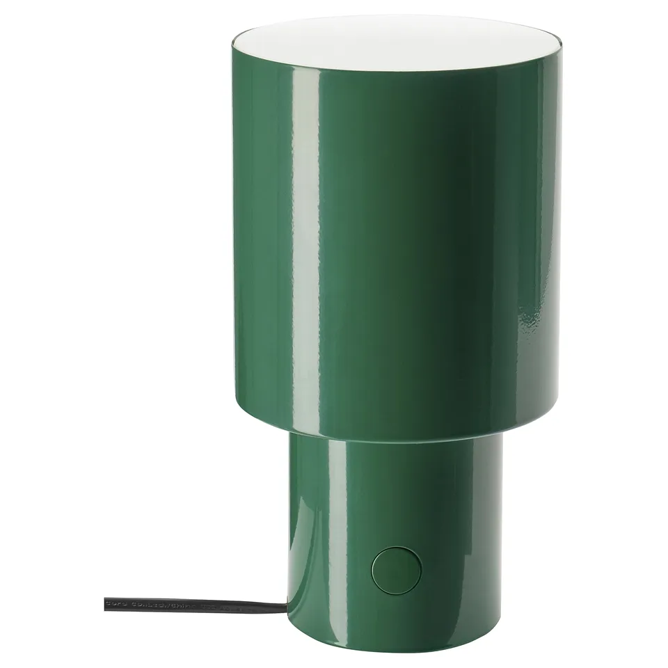 A green SPETSBOJ table lamp. It is cylindrical with a glossy finish and a round button on the side for control. The sleek design adds a modern touch.