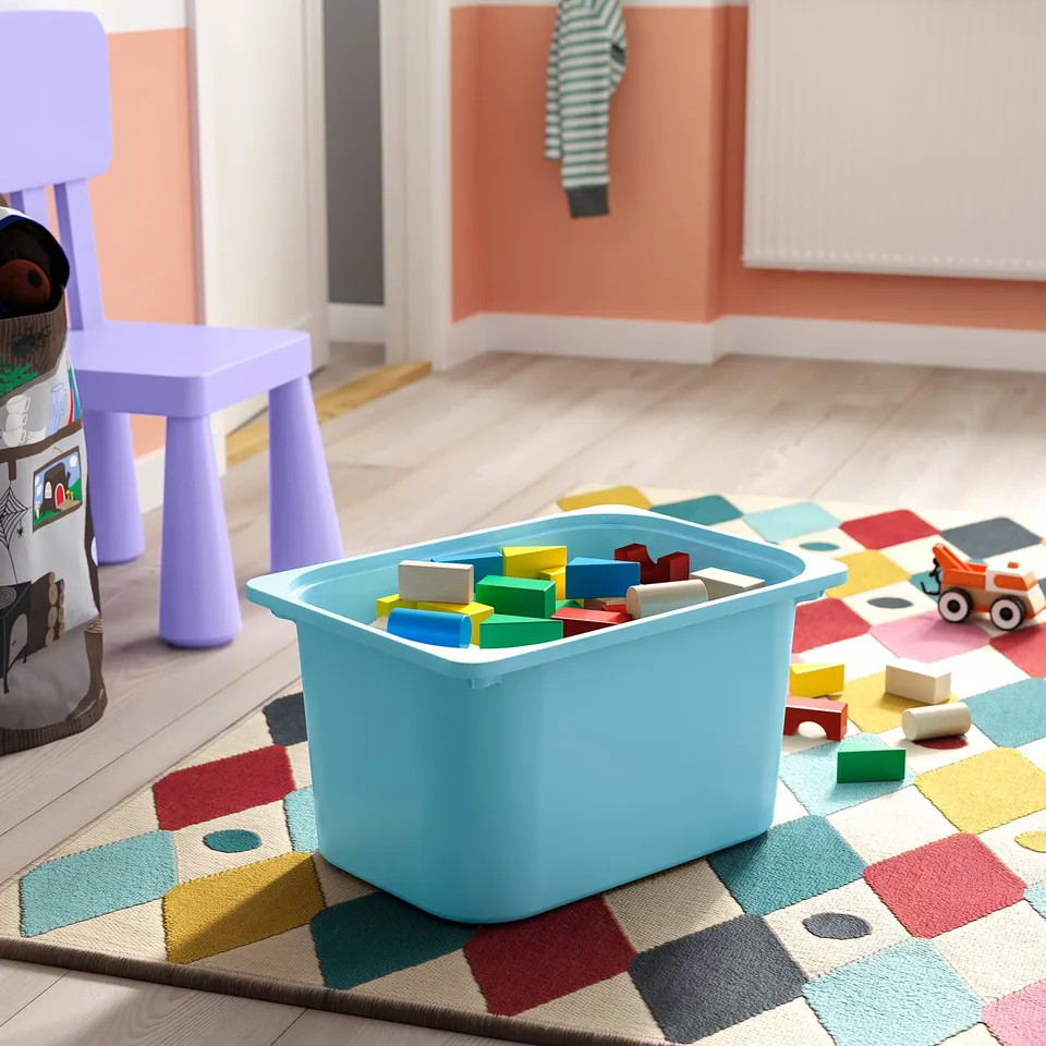 Light blue TROFAST storage bin filled with colourful wooden blocks sits on a multicoloured chequered rug in a childrens playroom.
