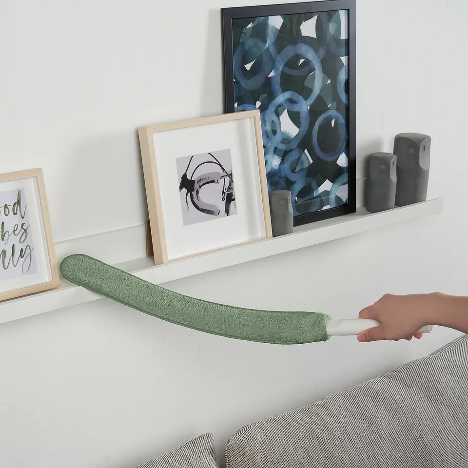 Hand uses PEPPRIG cleaning tool with microfiber pad, removing dirt under bed, against white wall, beside picture frames and shelves.