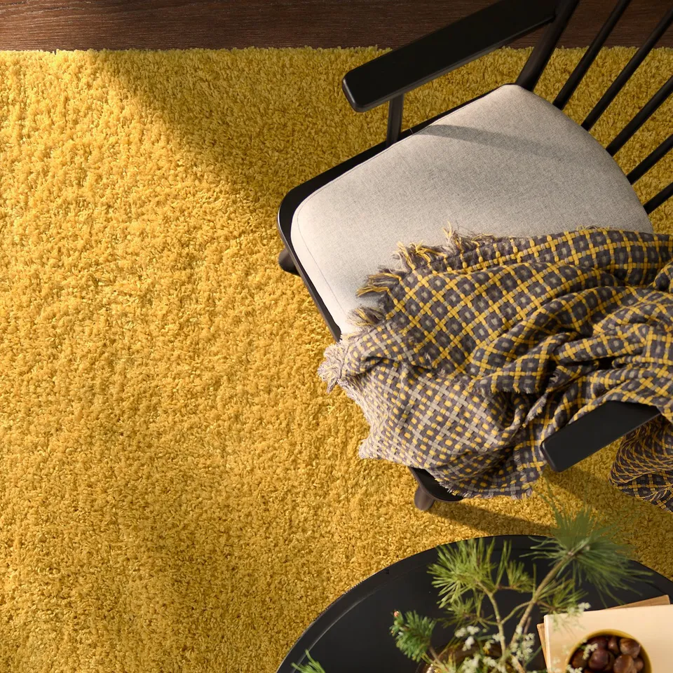 Mustard-colored ÄRENDE rug in living room with black chair & table, plant, yellow scarf.