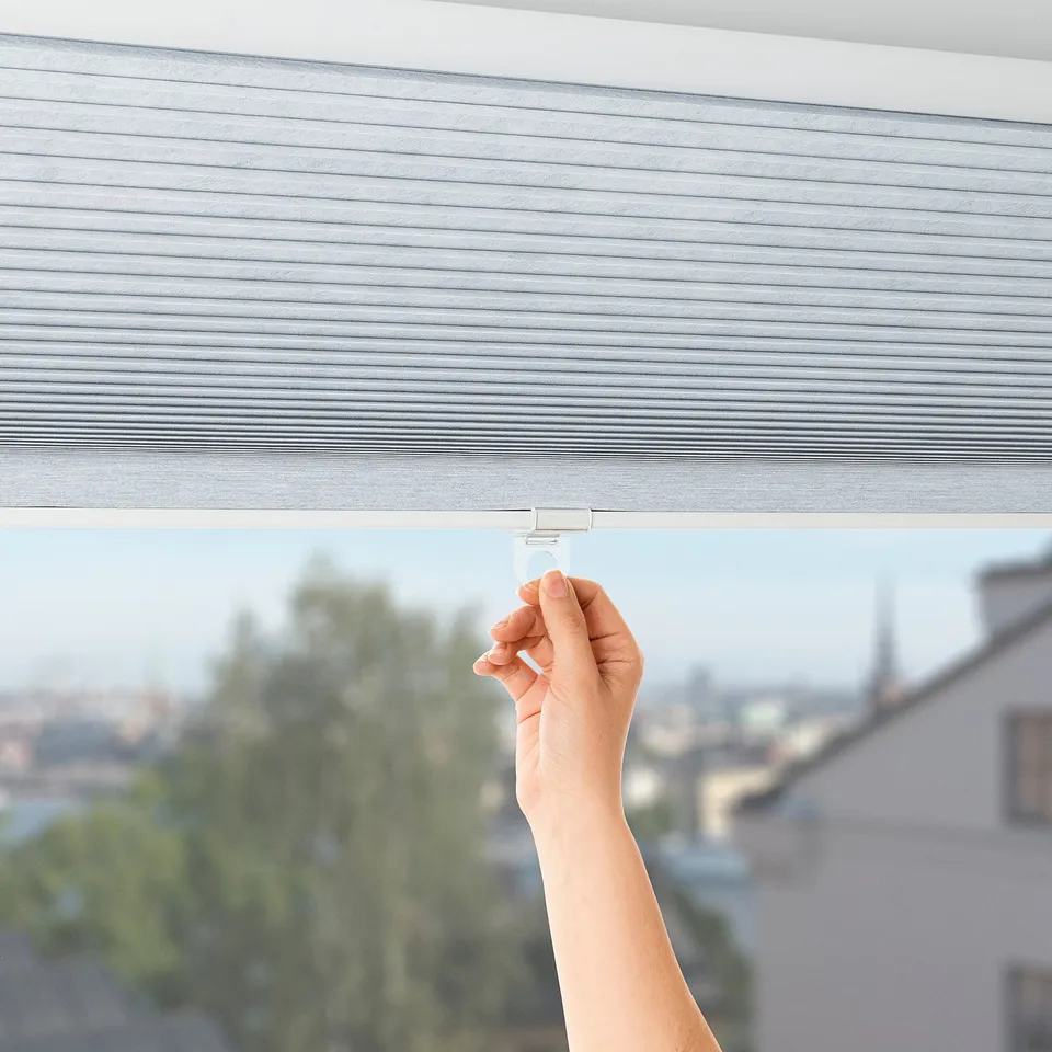 Hand lifting TRIPPEVALS block-out blind, revealing city view, demonstrating light control and child-safe design.