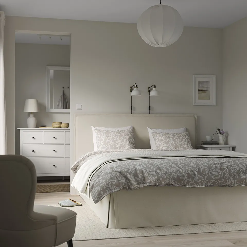 Calm bedroom with grey bed, patterned bedding, two night lamps, white dresser, and grey chair; bed is large yet fits well in room.