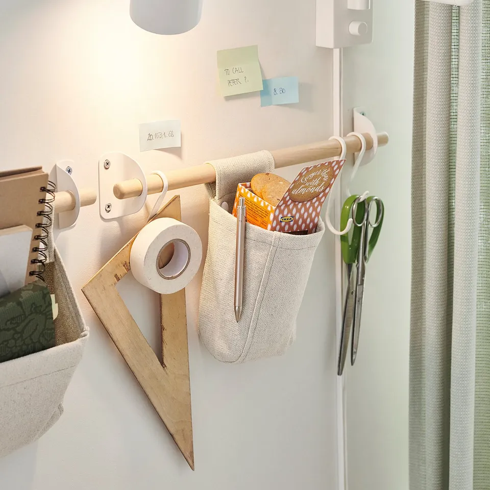 A white NEREBY wall rail system holds office supplies and notes. It includes hooks, containers, a lamp, and hanging items.