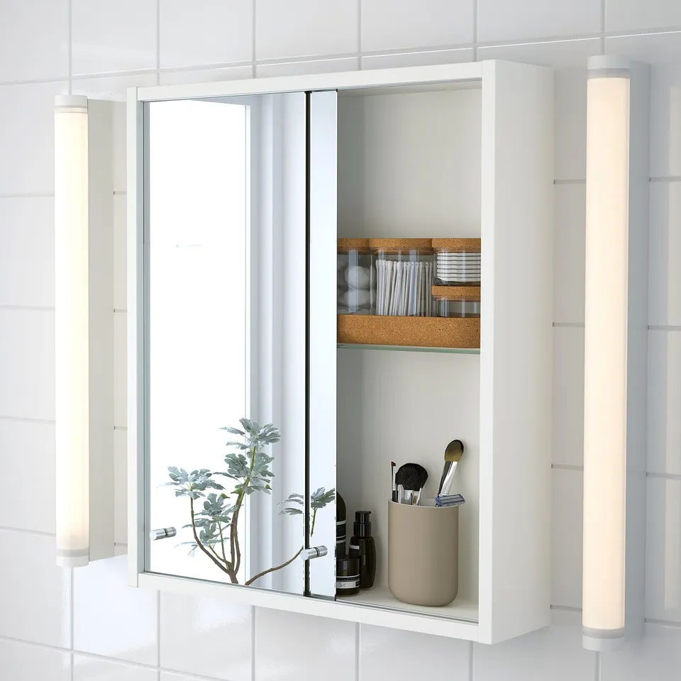Modern white cabinet with mirror, sliding doors, making contents accessible. Shelves hold makeup and a plant.