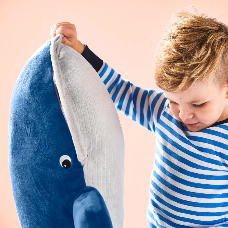 Child in striped shirt holds a large blue whale cuddly toy by the mouth, designed for playing and hugging.
