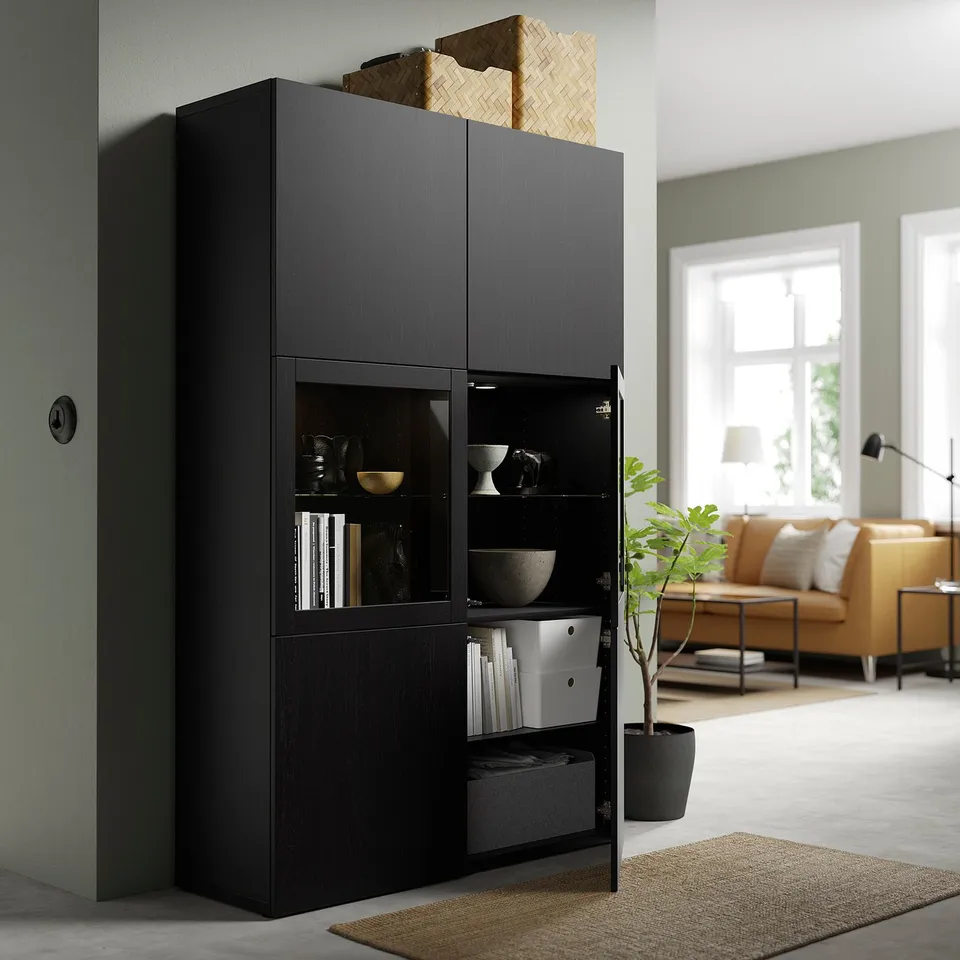 Black BESTÅ storage unit with glass doors, showcasing items, and adjustable shelves. Its placed in a living room with a couch and a window.
