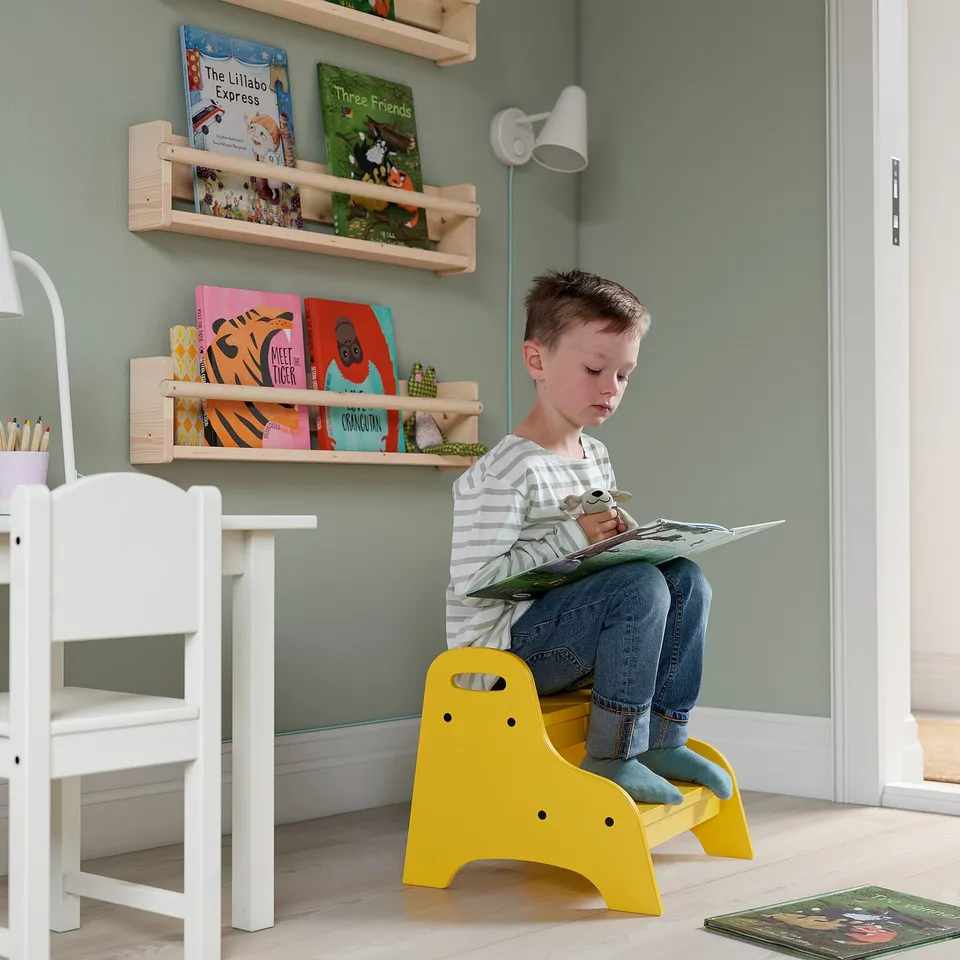 A young boy sits on a bright yellow TROGEN stool, reading a book in a cosy corner with bookshelves and a white chair beside the person.