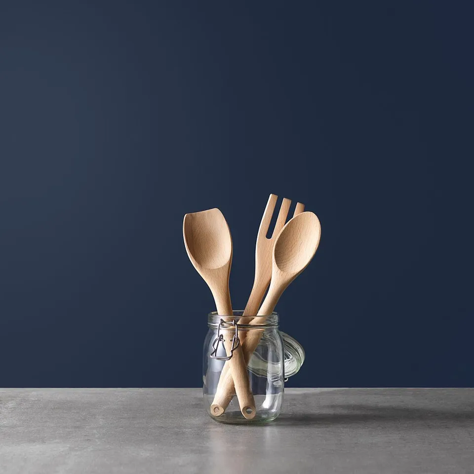 Three wooden cooking utensils in clear glass jar on grey counter, dark blue wall.