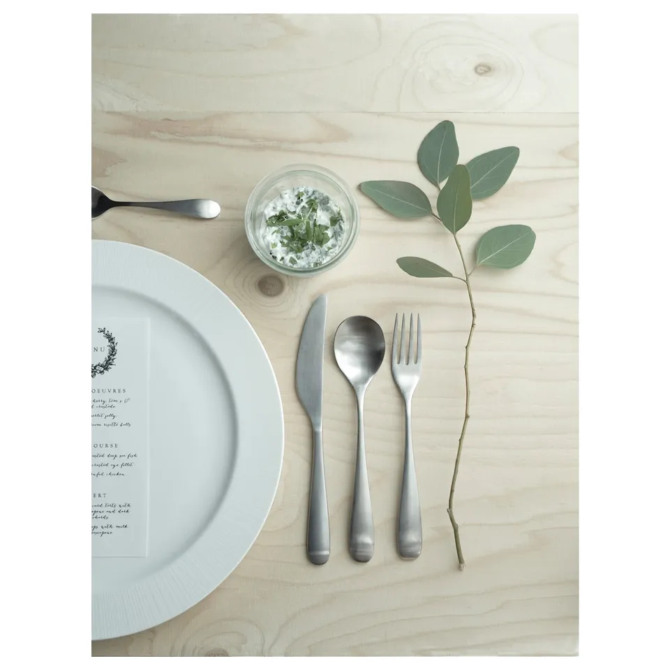 A table set with BEHAGFULL silver cutlery, a white plate, menu card, and green leaf branch on light wood.