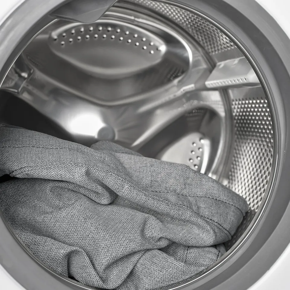 A washing machine drum with a grey fabric item inside. The drum has metallic perforated walls and a central agitator.