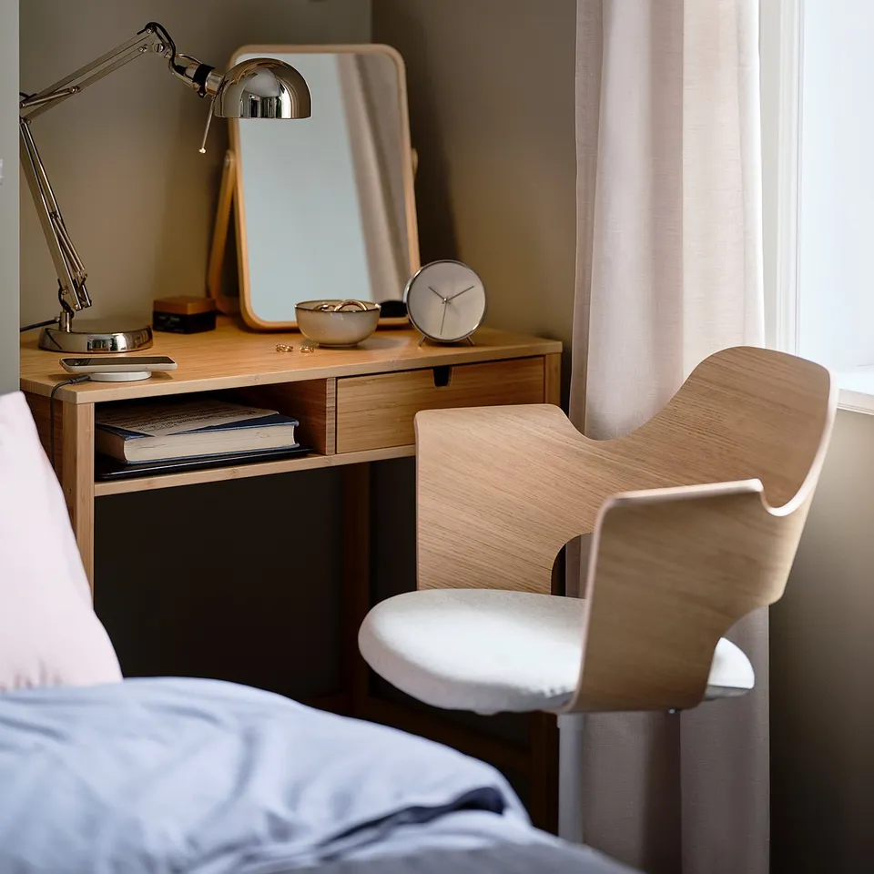 Modern bedroom workspace with light wood desk, white chair, adjustable silver desk lamp, square mirror, round clock, and curtains.