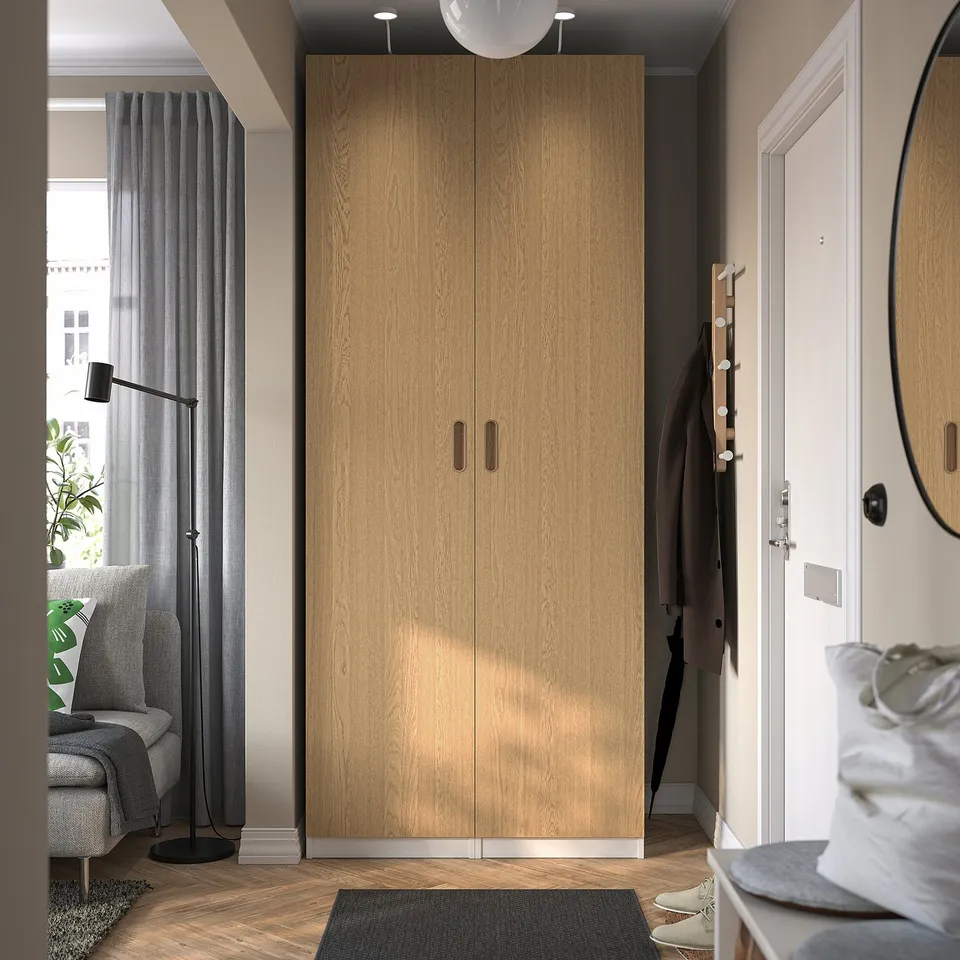 Large wooden TONSTAD wardrobe with handles, positioned between doorway and window, taller than surrounding decor items.