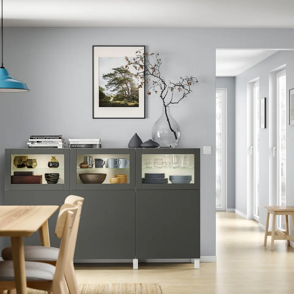Modern dining area with grey BESTÅ storage cabinet, glass doors, wooden table, and chairs.
