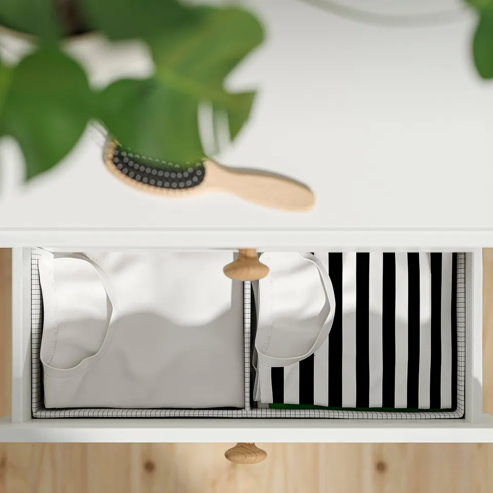 A minimalist chest of drawers with automatic drawer-closing system, holding white items, under a green plant.