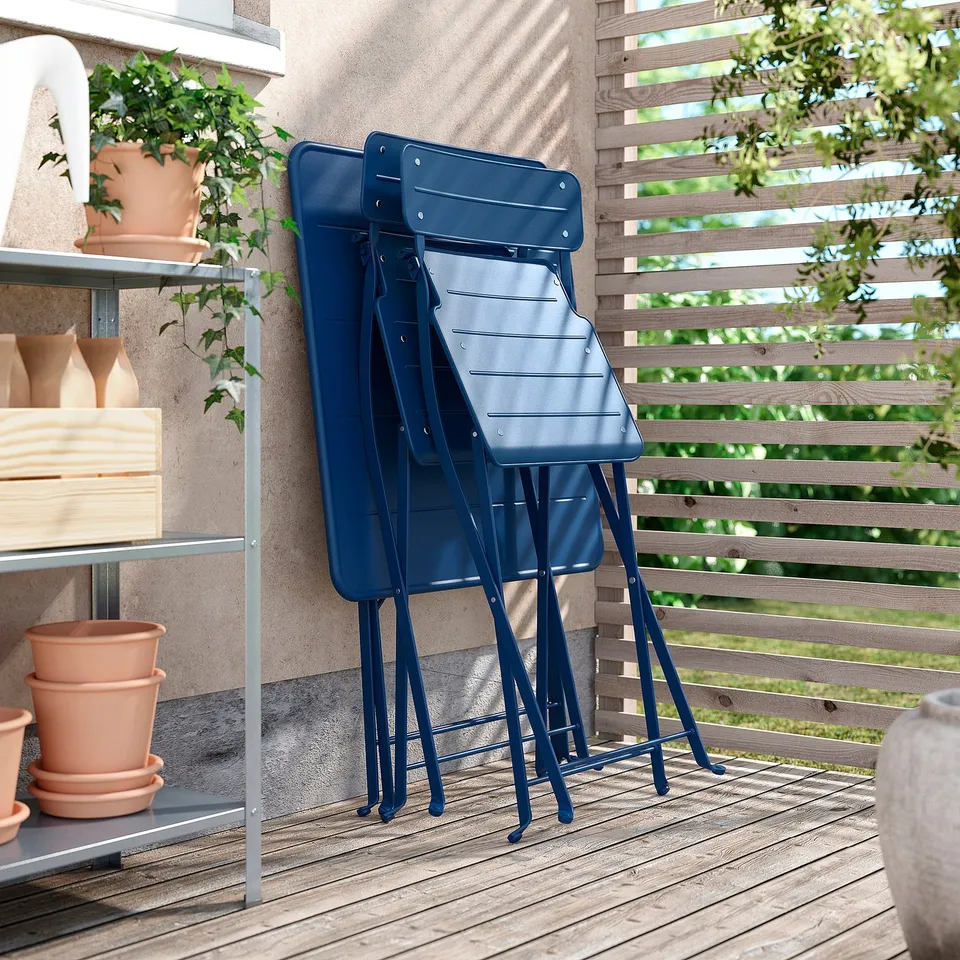 Blue metal chairs stacked neatly on wooden deck, leaning against wooden fence, beside outdoor shelving with terra cotta pots.