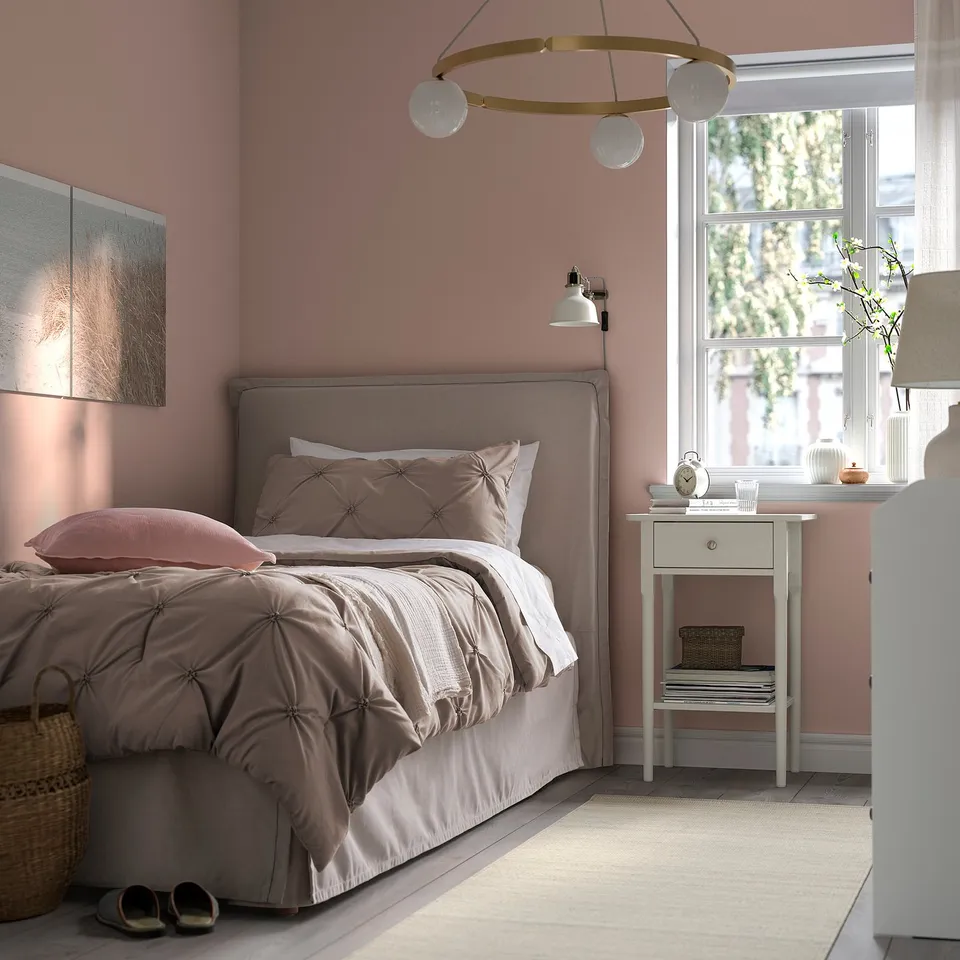 Cosy bed with BÄGARKLOCKA grey comforter, pillow shams, pink wall, bedside table, lamp, books, clock, and natural light.
