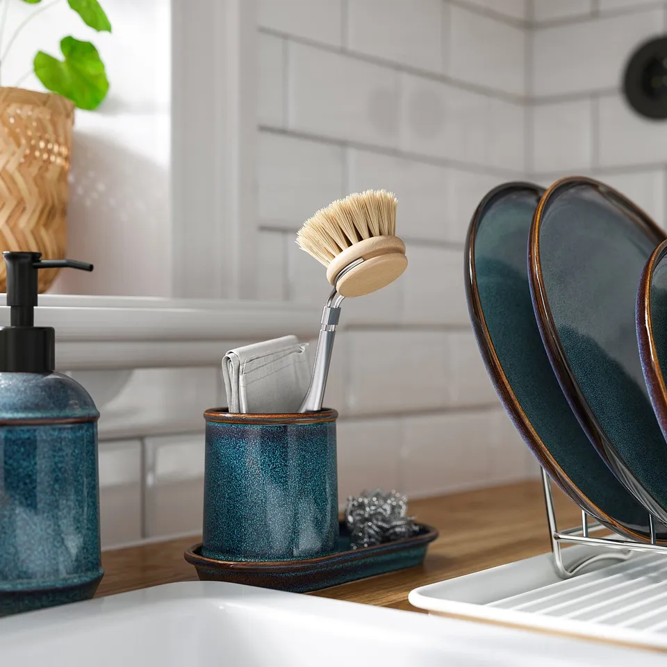 Kitchen sink with blue soap dispenser, toothbrush holder, and ceramic tray storing brushes and scouring pad. Tray holds small items.