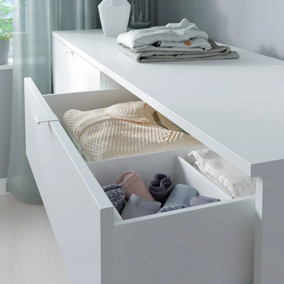 White drawer chest with folded clothes. Neatly organised light grey and beige textiles. Minimalist design for bedroom or living room.