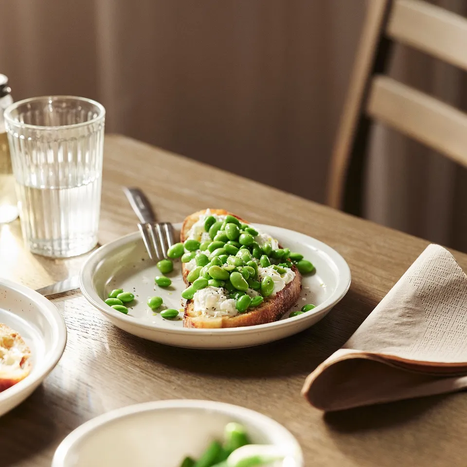 A rustic plate with toast topped with green peas and sauce, on a wooden table with a glass, fork, and napkin.