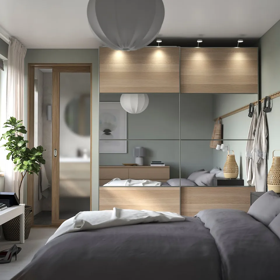 Modern bedroom with PAX wardrobe, mirrored sliding doors, grey lights, bedding, plant, minimalist decor.