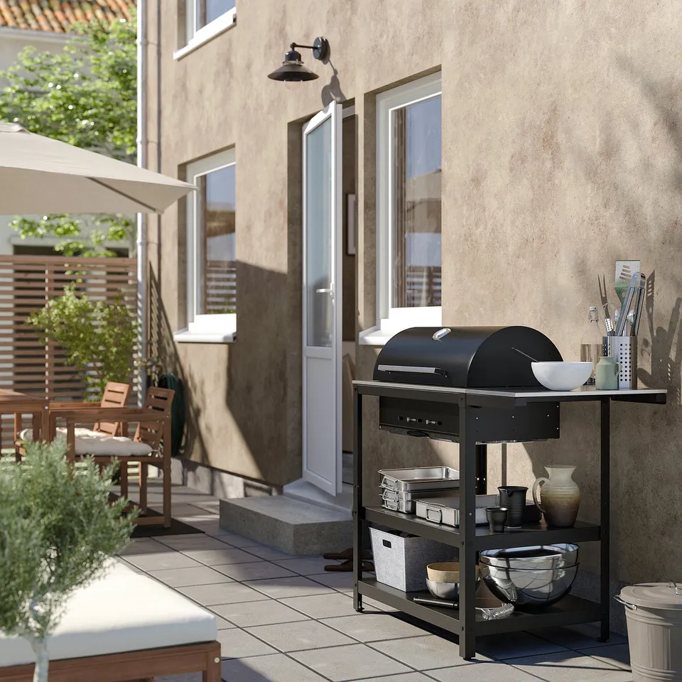 Black outdoor grill station, stainless steel top with tools, beside house entrance, beige wall background, tiled patio.