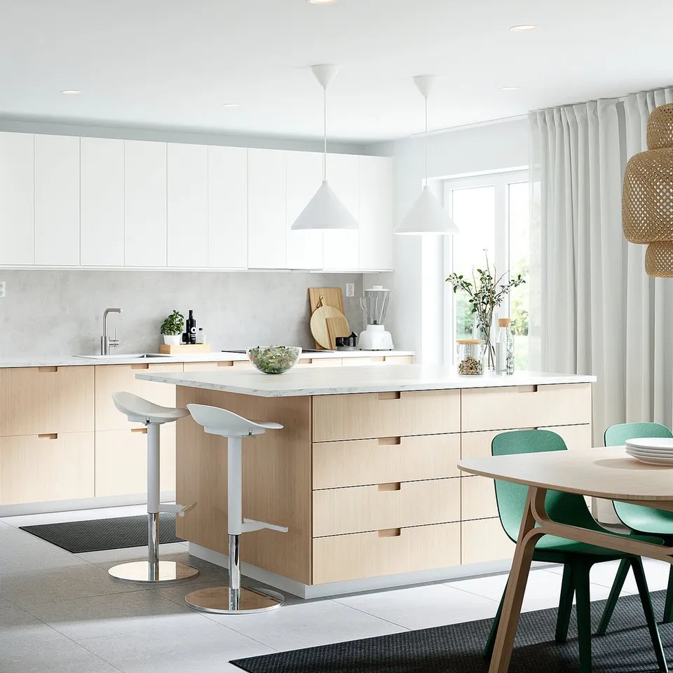 Modern white kitchen with light wood island, white barstools, green dining chairs, hanging lamps, and numerous potted plants.