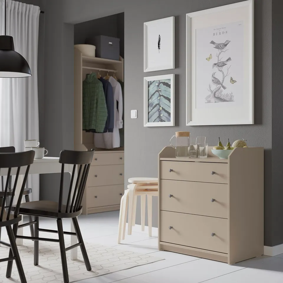 Modern living space with HAUGA light beige chest, art prints, wooden chairs, and open closet.