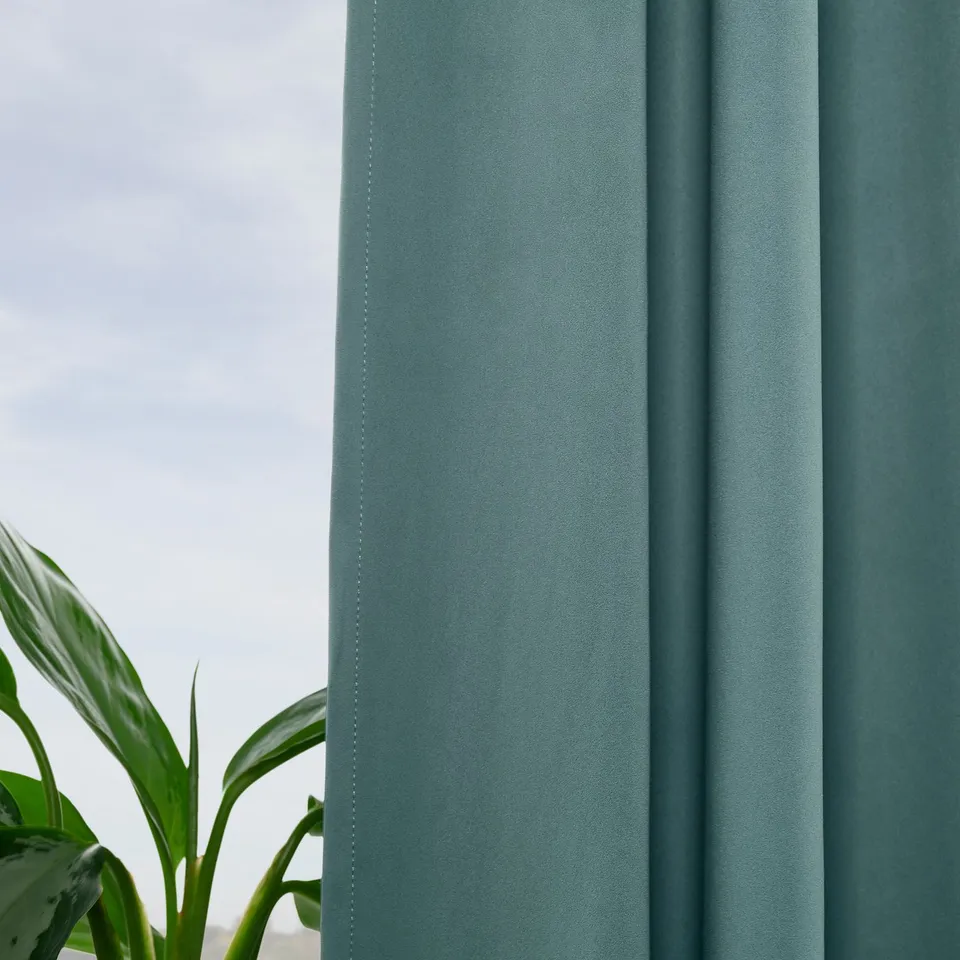Teal room-darkening curtains hang beside a green plant, offering privacy and temperature control.