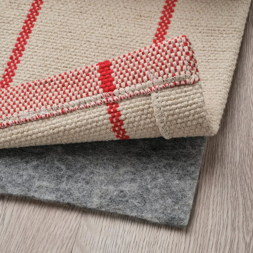 Textured beige rug with red stripes folded atop black non-slip pad on wooden floor.