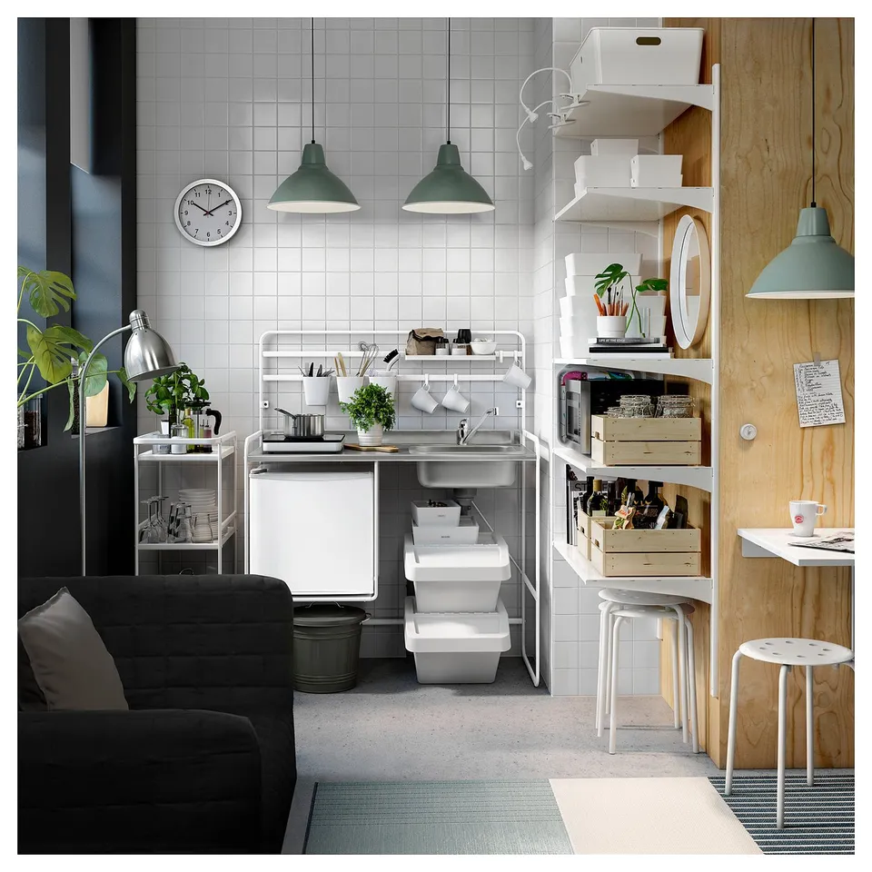 Compact modern kitchen with tiled walls, hanging lamps, and open storage. White and wood elements, a black sofa, and a stainless-steel sink.