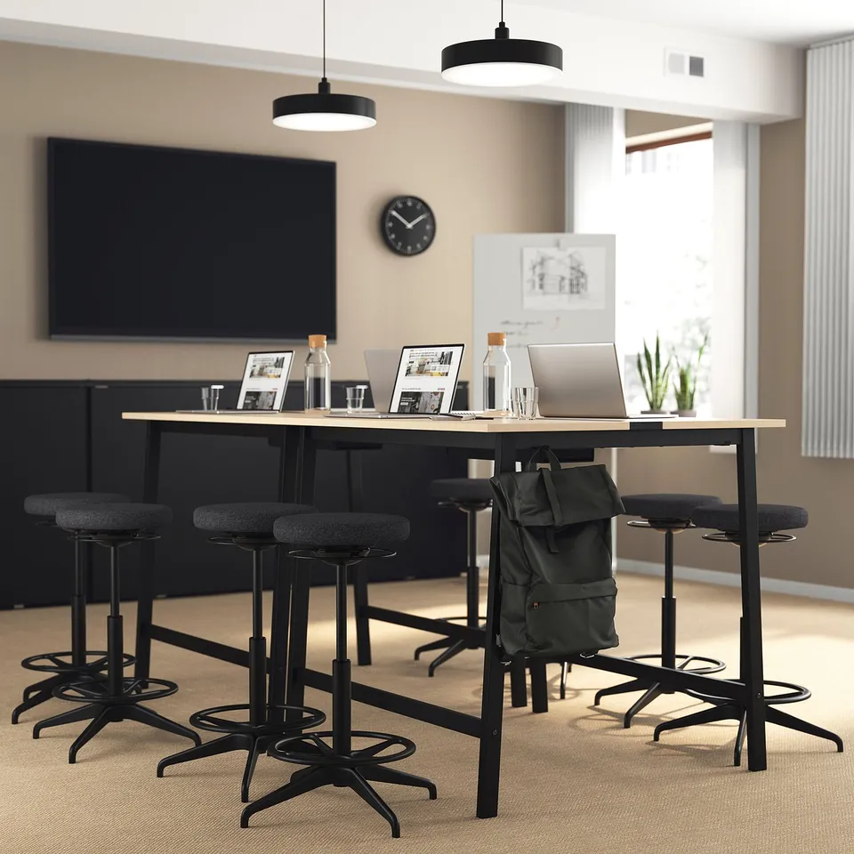 Modern office with MITTZON high table, screen, plants. Table has laptop, bottles, frames. Small round stools.