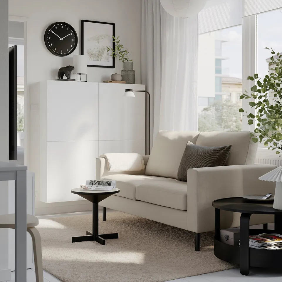 Modern living room with beige SALTSJÖBADEN sofa, black coffee & side tables, white cabinet with clock, tall plant, minimalist design.