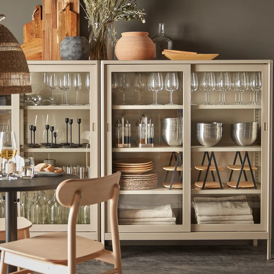 Modern cream-colored IDÅSEN cabinet with glass doors, housing wine glasses, bowls, plates, and candles. Wooden chair beside it.