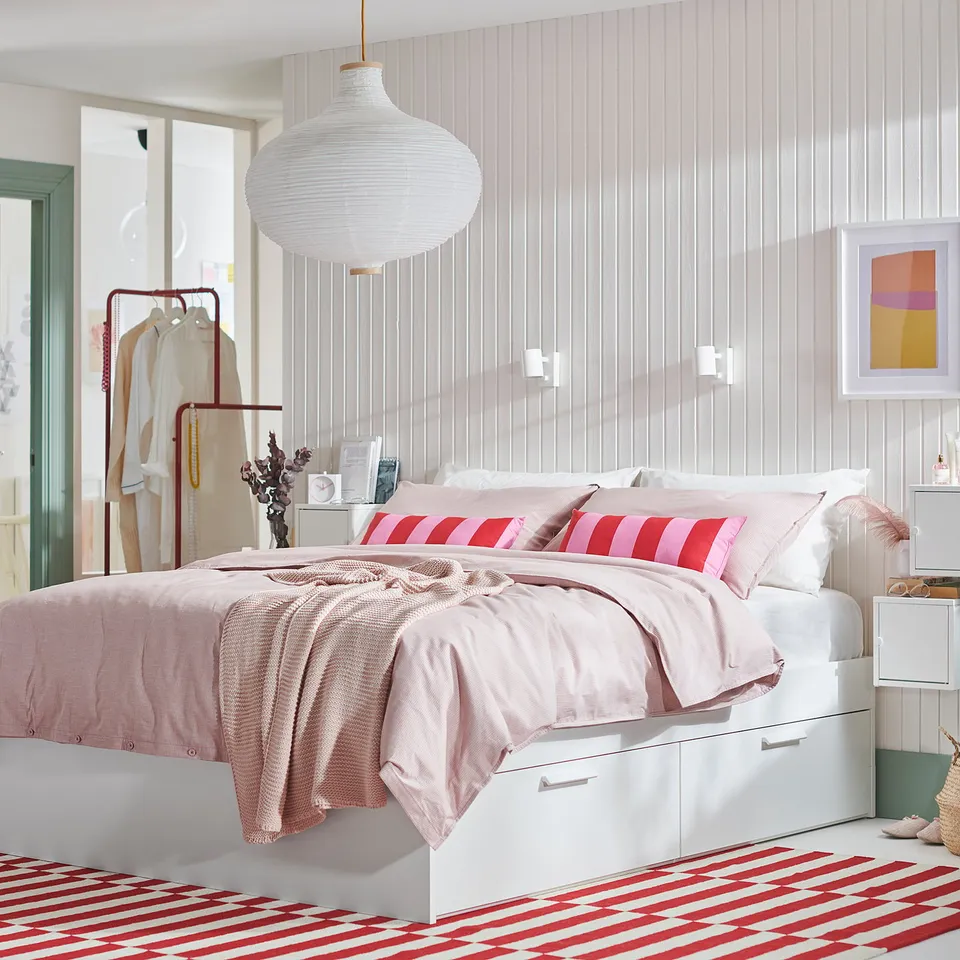Bedroom with white BRIMNES bed, light pink linens, red-white rug, hanging light, white walls, striped wallpaper, and framed art.