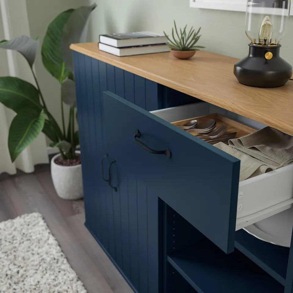 Navy blue sideboard with open drawer revealing cutlery, topped with wooden surface holding books, a potted plant, and a lamp.
