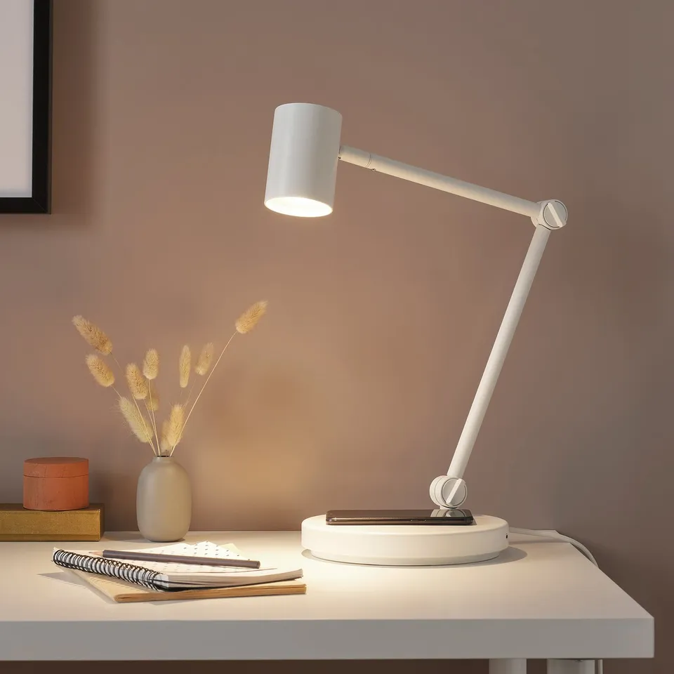 NYMÅNE white desk lamp with adjustable arm on a white desk. Next to a vase with dried flowers and notepad. Modern, minimalist design.